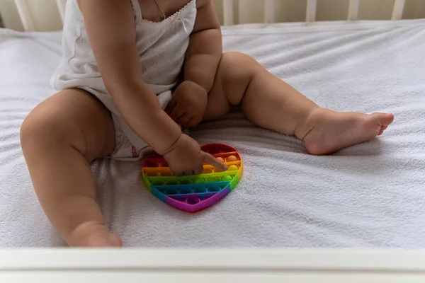 可爱的幼儿小女孩坐在那里玩彩虹爸爸它的手指玩具在白色的婴儿床在白色的夏天睡衣和枕头在家里。童年、闲暇、舒适、发展概念 — 图库照片