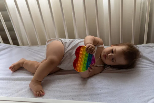 Симпатичная маленькая девочка, сидящая дома в белой кроватке в белой летней пижаме с подушкой, играет в Rainbow Pop. Детство, отдых, комфорт, концепция развития — стоковое фото