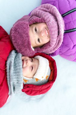 Çocukluk, karlı zamanlar, kış tatilleri, soğuk hava. Anaokulunun küçük mutlu portresi, kız çocuk yüzü, kalın örülü şapka ve eşarp takmış, beyaz kar arka planına uzanmış çocuklar.