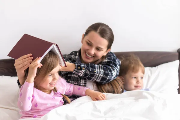 Glückliche Familie. Nahaufnahme liebevolle Mutter liegend mit Tochter Sohn zwei Kinder Pyjama im Bett Kinder lesen interessante Bilderbuch, Vorschule lächelnde Kind Mädchen genießen Märchen vor dem Schlafengehen Buch auf dem Kopf — Stockfoto