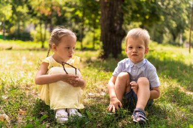Mutlu gülümseyen iki çocuk anaokulu ikizi iki kardeş kardeş kardeş kardeş kardeş kız kardeş güneşli yaz havasında parkta çimlerin üzerinde birlikte oturuyorlar. çocukluk, arkadaşlık, aile kavramı