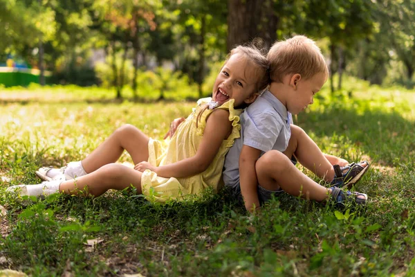 Две счастливые улыбающиеся веселые детские дошкольники-близнецы братья и сёстры сидят вместе на газоне в парке в солнечную жаркую летнюю погоду. детство, дружба, понятие семьи Стоковая Картинка