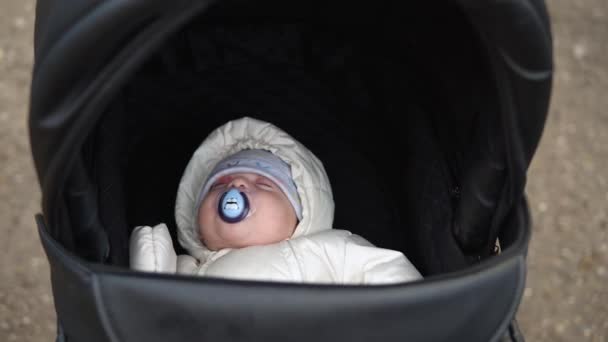 Παιδική ηλικία, βρεφική ηλικία, χειμώνας. close-up Μικρό μωρό κοιμάται σε ζεστό κάτω σακάκι σε μαύρο καρότσι. Η μαμά περπατάει ξάγρυπνο νήπιο βρέφος σε καροτσάκι στην αυλή με κρύο φθινοπωρινό καιρό έξω στο πάρκο. — Αρχείο Βίντεο