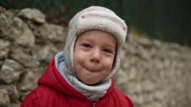 Gri örgülü kırmızı ceketli, kameraya gülümseyen küçük Avrupalı anaokulu çocuğunun yakın plan portresi. Duygusal çocuk gülüyor, gülümsüyor, elleriyle gözleri örtüyor, sonbaharın dışında el sallıyor.