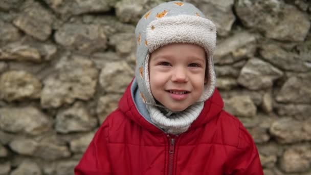 Spowolniony ruch zbliżenie Portret europejskiego małego przedszkolaka w szarej dzianinie kapelusz czerwona kurtka uśmiechnięta spojrzeć w aparat fotograficzny. Emocjonalnie dziecko śmiejące się uśmiechnięte, pozujące oczami z okładki z rękami na zewnątrz Jesień — Wideo stockowe