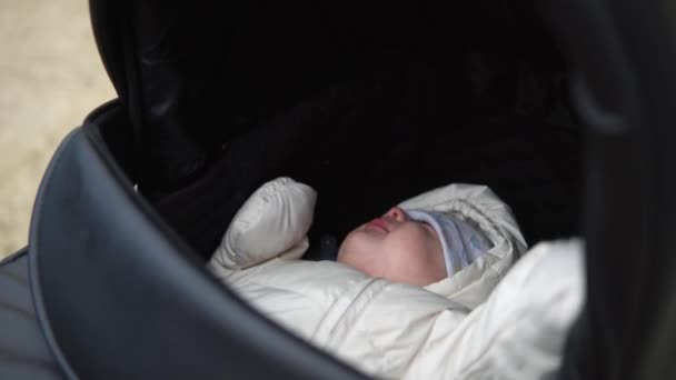 Infancia, infancia, invierno. primer plano Pequeño bebé dormir en chaqueta de abrigo en cochecito negro. Mamá camina tranquilamente niño dormido bebé en cochecito en el patio en el frío clima de otoño fuera en el parque de carreteras — Vídeos de Stock