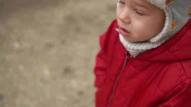 Gri örgülü kırmızı ceketli, kameraya gülümseyen küçük Avrupalı anaokulu çocuğunun yakın plan portresi. Duygusal olarak çocuk üzgün rüya poz veriyor ellerle gözleri örtüyor, sonbaharın dışında el sallıyor merhaba