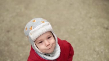 Gri örgülü kırmızı ceketli, kameraya gülümseyen küçük Avrupalı anaokulu çocuğunun yakın plan portresi. Duygusal olarak çocuk üzgün rüya poz veriyor ellerle gözleri örtüyor, sonbaharın dışında el sallıyor merhaba