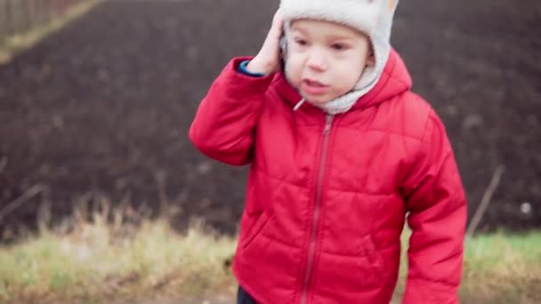 Närbild Porträtt av europeiska lilla förskolekillen i grå stickad hatt röd jacka ler titta på kameran. Känslomässigt barn skrattar ler talar talar per telefon, vågor hej utanför hösten — Stockvideo