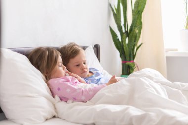 Küçük anaokulu çocuğu küçük çocuklar, çocuklar çizgi film izlerken birlikte akıllı telefon cihazı kullanırlar. Evde, beyaz yatakta pijamalı bebek var. Aile, Boş Zaman, Çocukluk, Arkadaşlık Konsepti