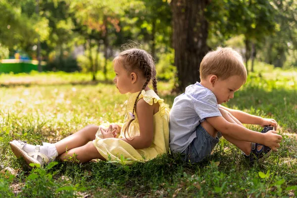 Две счастливые улыбающиеся веселые детские дошкольники-близнецы братья и сёстры сидят вместе на газоне в парке в солнечную жаркую летнюю погоду. детство, дружба, понятие семьи — стоковое фото