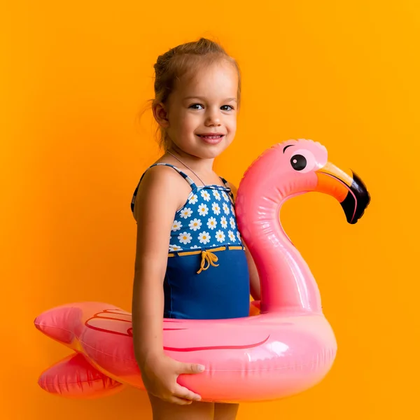 Счастливая маленькая девочка-пловчиха носит солнечные очки купальник, ребенок надувной плавательное кольцо Розовый Фламинго указывают в сторону, изолированный оранжевый фон. детство, жаркие летние каникулы Стоковое Фото