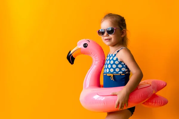 Šťastný malý plavec sportovní dívka nosit sluneční brýle plavky, Kid Nafukovací plavecký kroužek Pink Flamingo bod na stranu, izolované oranžové pozadí. dětství, horká letní dovolená koníček koncept — Stock fotografie