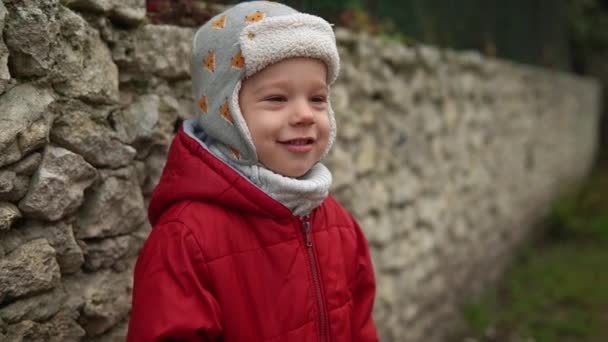 Close-up Portrait of European little preschool boy in gray knitted hat red jacket smiling look at camera. Emotionally child laughing smiling posing cover eyes with hands, waves hello outside Autumn — Stock Video