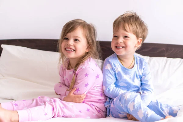 Двое детей дошкольного возраста, мальчик и девочка улыбаются, смотрят на сторону в розово-голубой пижаме на белой кровати. Малыши-близнецы веселятся. Счастливые дети на карантине дома. Дружба, семейная концепция — стоковое фото