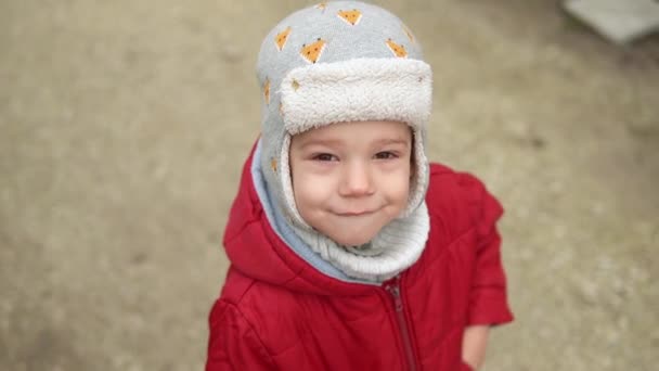 Närbild Porträtt av europeiska lilla förskolekillen i grå stickad hatt röd jacka leende titta på kameran. Känslomässigt barn skrattar poserar promenader slow motion, vågor hej utanför hösten — Stockvideo