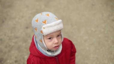 Gri örgülü kırmızı ceketli, kameraya gülümseyen küçük Avrupalı anaokulu çocuğunun yakın plan portresi. Duygusal olarak çocuk üzgün rüya poz veriyor ellerle gözleri örtüyor, sonbaharın dışında el sallıyor merhaba