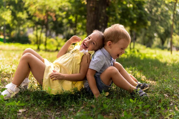 Две счастливые улыбающиеся веселые детские дошкольники-близнецы братья и сёстры сидят вместе на газоне в парке в солнечную жаркую летнюю погоду. детство, дружба, понятие семьи — стоковое фото
