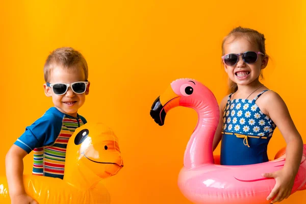 Dvě šťastné malé děti plavec sportovní dívka chlapec nosí sluneční brýle plavky, Kid Nafukovací plavecký kroužek ze strany na stranu, izolované oranžové pozadí. dětství, horká letní dovolená koníček koncept — Stock fotografie