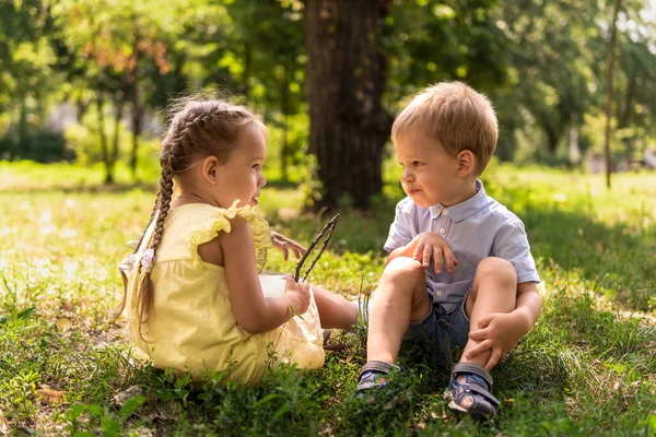 Две счастливые улыбающиеся веселые детские дошкольники близнецы близнецы братья и сёстры девочки общаются друг с другом на газоне в парке в солнечное жаркое лето. детство, дружба, понятие семьи — стоковое фото