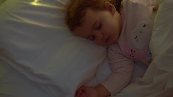 Linda niña Chuby 1-2 años durmiendo en pijama rosa en cama blanca en dormitorio oscuro. Niño tener descanso tiempo en la noche guirnalda en la almohada. Infancia, Navidad, Concepto de paternidad — Vídeo de stock