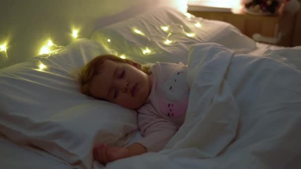 Roztomilé Chuby Malá batole Baby Girl 1-2 Let staré spí v růžové pyžamo na bílé posteli v tmavé ložnici. Dítě Odpočiňte si v noci Garland na polštáři. Dětství, Vánoce, Koncept rodičovství — Stock video