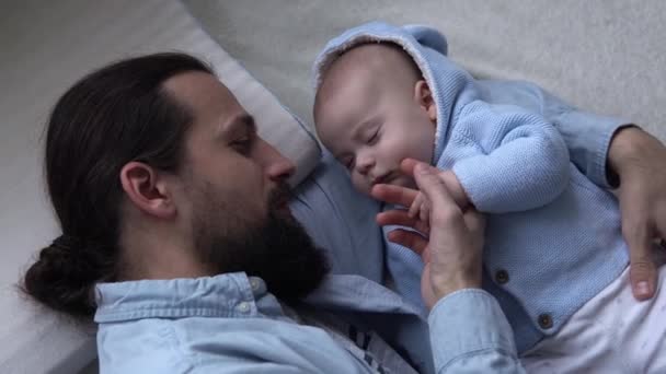 Cinematic Shot Bearded Long-haired Young Neo Father Strokes Face Newborn Baby In Nursery At Home. Dad Laying With Infant Child. Children, Parenthood, Childhood, Life, Love, Fatherhood, Family Concept — Vídeos de Stock