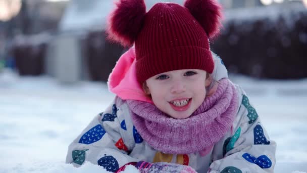 Close up Portrait Little Child Pretty Girl Kid Smiling Looking at Camera play red hat lie on Stomack. Snow Park Outdoor. Winter time, happiness concept Snowing Cold frosty weather Carefree childhood — Wideo stockowe