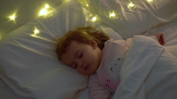 Söt Chuby Little Toddler Baby Girl 1-2 år gammal sovande i rosa Pajama på vit säng i mörkt sovrum. Barn har koppla av vilotid på natten Garland på kudde. Barndom, Jul, Föräldraskap Begreppet — Stockvideo