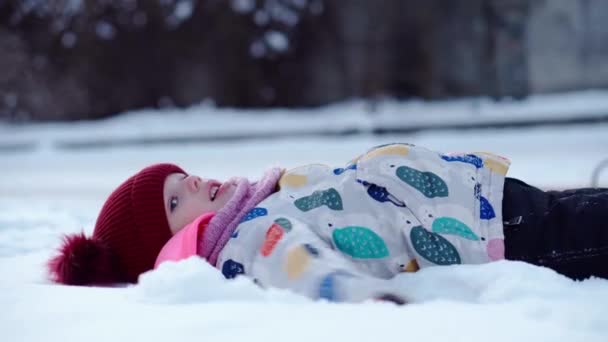 Close up Portret klein kind mooi meisje kind glimlachend kijkend naar camera spelen in rode hoed liggen op de rug. Sneeuwpark Buiten. Wintertijd, geluksconcept. Koud ijskoud weer. Zorgeloze jeugd — Stockvideo