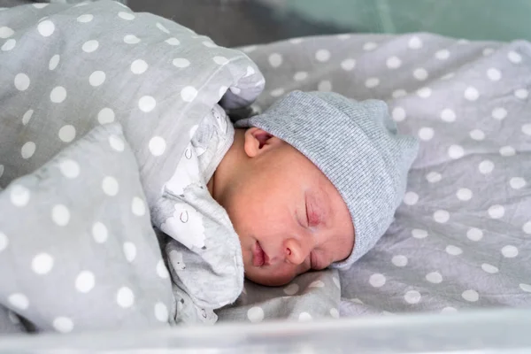 Recém-nascido bebê vermelho bonito rosto retrato primeiros dias dormindo na cama de vidro médica em fundo cinza. Criança no início minutos de vida no chapéu. Bebês, Parto, Primeiros Momentos de Borning, Princípio de Início — Fotografia de Stock