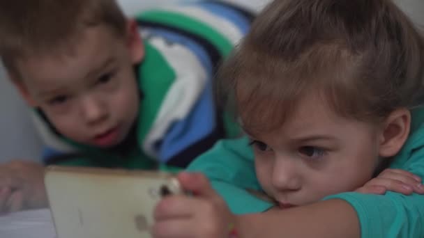 Auténticos 3 niños pequeños preescolares niños menores hermanos ven dibujos animados en el dispositivo del teléfono inteligente juntos bebé en la cama blanca en casa en interiores. Familia, ocio, infancia, concepto de amistad. — Vídeos de Stock