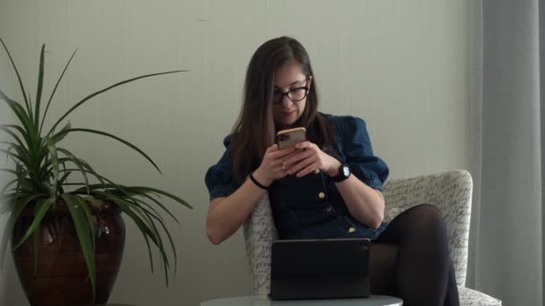 Успешная молодая женщина в очках болтая по телефону гостиная. Писать с помощью информационных технологий. Счастливая улыбающаяся барышня работает в Интернете. Образование, технологии — стоковое видео