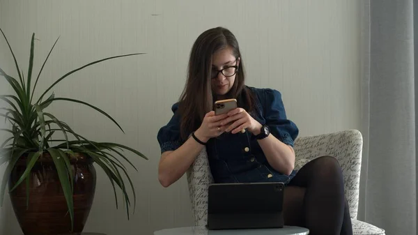Gözlüklü başarılı bir genç kadın. Dizüstü bilgisayarın yanında sohbet ediyor. Kullanarak Yazma Araması. Smartphone Browsing Internet 'te çalışan Mutlu Gülümseyen Bayan Buisenes. Eğitim, Teknoloji Konsepti - Stok İmaj