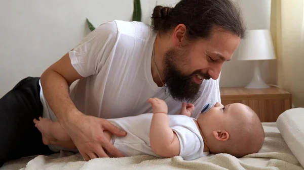 Homem Barbudo Jovem Autêntico Segurando Bebê Recém-nascido. Pai e filho na cama. Close-up Retrato de família sorridente com o bebê nas mãos. Casamento feliz Casal no fundo. Infância, Conceito de paternidade — Fotografia de Stock