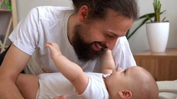 Homem Barbudo Jovem Autêntico Segurando Bebê Recém-nascido. Pai e filho na cama. Close-up Retrato de família sorridente com o bebê nas mãos. Casamento feliz Casal no fundo. Infância, Conceito de paternidade — Fotografia de Stock