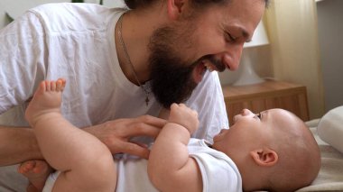 Otantik Sakallı Genç Adam Yeni Doğan Bebeği Tutuyor. Baba ve çocuk yataktalar. Ellerinde Bebekli Gülümseyen Aile portresi. Arka plandaki mutlu evlilik çifti. Çocukluk, Ebeveynlik Konsepti