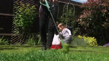 Çim biçme makinesi çim biçiyor. Çim biçme makinesinden çıkan küçük yeşil çim biçme makineleri peyzajcılar tarafından itilip kakılıyor. Ağır çekim. Bahçıvan adam bahçe bahçesinde çim biçme makinesiyle çalışıyor. Aile, iş..