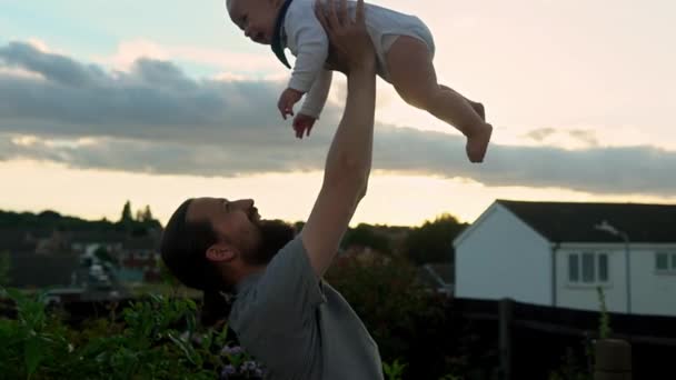 快乐的父亲抱着可爱的男婴拥抱亲吻 笑爸爸和儿子在花园外面玩 日落时的全家福 爸爸飞起来的孩子就像飞机一样快乐 父母和孩子在一起很开心 — 图库视频影像