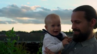 Happy Father holding adorable baby boy embracing kissing. Smiling Dad with son playing outside Garden. Family silhouette at sunset. Daddy fly up child as airplane having fun. Parent enjoying with kid.