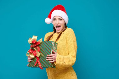 Noel Baba şapkalı mutlu genç kadın elinde yeni yıl hediyesi ve mavi izole edilmiş arka planda gülüyor.