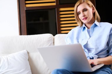 Günlük kıyafetli bir kadın dizüstü bilgisayarda çalışıyor. Evdeki kanepede oturuyor..