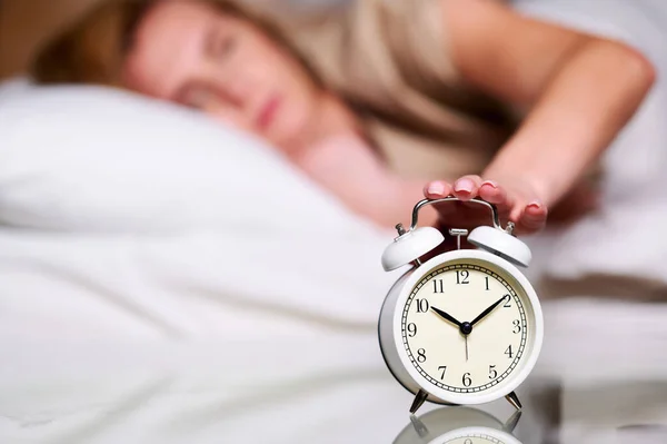 Kapatmak için çalan alarma elini uzatan uykulu beyaz bir kadın. Erken kalkmak, uykusuzluk, iş kavramı.. Stok Fotoğraf