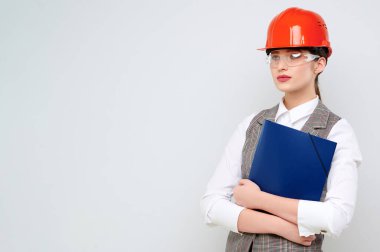 Young female caucasian engineer wearing orange constraction helmet and googles holds blueprint on isolate background