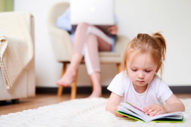 Girl reads a book while lying on the floor while mom works on a laptop at home while sitting on an armchair