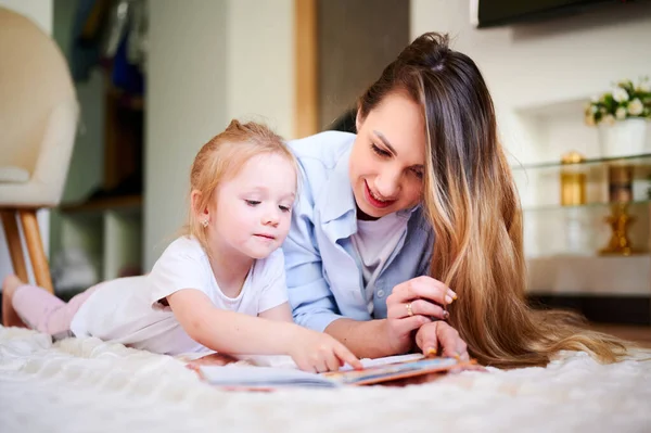 Jeune mère avec sa jolie petite fille lit le livre et regarde les dessins dans le livre tout en étant couché sur le sol. La mère enseigne à l'enfant. La relation entre parents et enfants — Photo