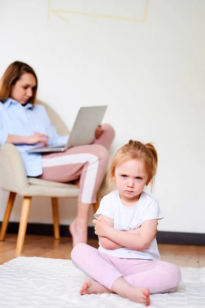 Rozrušená holčička sedící osamělá, zatímco matka používá notebook. Matka nemá čas na dceru. — Stock fotografie