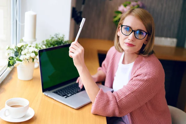 Joven hermosa mujer caucásica en gafas trabaja en un ordenador portátil tiene una pluma en sus manos bebe café trabajo de oficina en casa durante la cuarentena — Foto de Stock