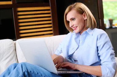 Günlük kıyafetli bir kadın dizüstü bilgisayarda çalışıyor. Evdeki kanepede oturuyor..