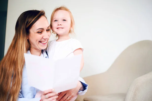 С Днем Матери. Счастливая улыбающаяся мама с маленькой дочкой читает праздничную открытку, сидя на полу — стоковое фото